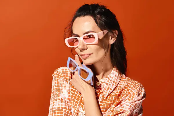 A fashionable woman poses in pink sunglasses and a colorful patterned shirt on a vibrant orange background. — Stock Photo
