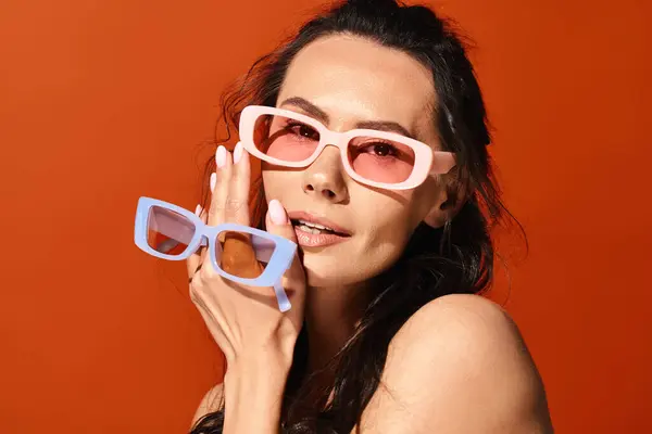 Une femme élégante dans des lunettes roses tient une paire de lunettes de soleil dans un contexte orange vif. — Photo de stock