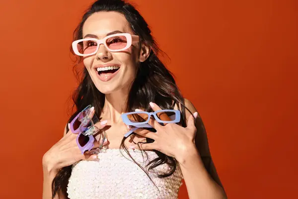 A stylish woman in a white dress and pink glasses poses gracefully in a studio with an orange background, embodying summertime fashion. — Stock Photo