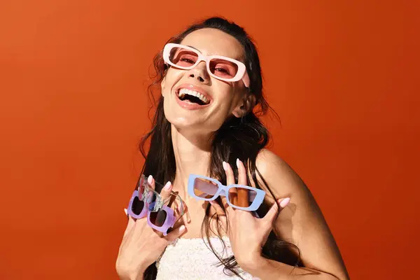 Una mujer impresionante exuda felicidad mientras sonríe hacia la cámara con elegantes gafas de sol, contra un vibrante fondo de estudio naranja. - foto de stock