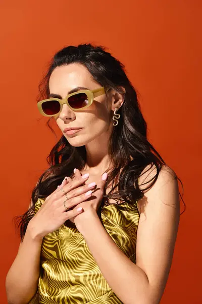 Une femme élégante dégage des vibrations estivales dans une robe jaune vibrante et des lunettes de soleil chics sur fond de studio orange. — Photo de stock