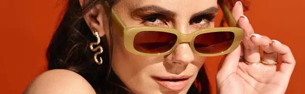 Une femme à la mode dégage des vibrations estivales dans un studio, berçant des lunettes de soleil jaune vif sur fond orange. — Photo de stock