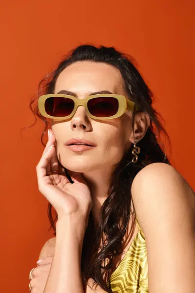Une femme élégante dans une robe jaune vibrante et des lunettes de soleil chics pose gracieusement sur un fond orange, respirant des vibrations estivales. — Photo de stock