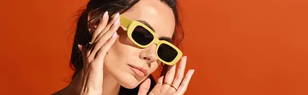 Une femme élégante portant des lunettes de soleil jaunes pose avec ses mains délicatement placées sur son visage, respirant la confiance et la mode estivale sur un fond de studio orange. — Photo de stock