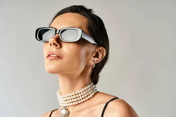 A stylish woman exudes sophistication as she wears sunglasses and a pearl necklace in a studio against a grey background. — Stock Photo