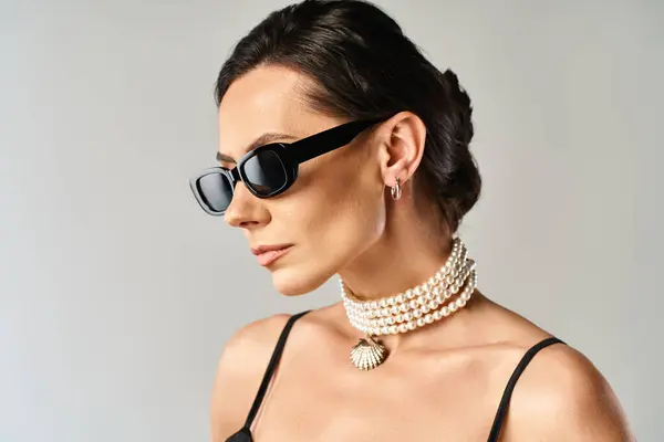 A fashionable woman with stylish sunglasses and a chic necklace poses in a studio against a grey background. — Stock Photo