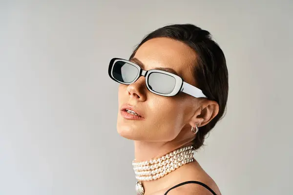 Uma mulher elegante com óculos de sol e um colar de pérolas transpira confiança em um estúdio contra um fundo cinza. — Fotografia de Stock