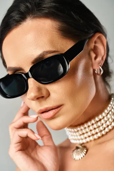 Une femme à la mode respire la sophistication dans les lunettes de soleil noires et un collier de perles sur fond neutre. — Photo de stock