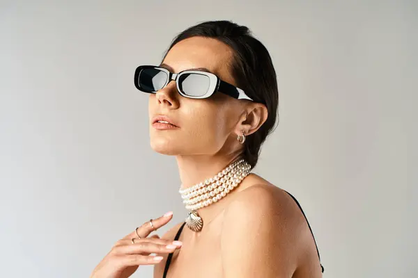 Una donna elegante con occhiali da sole e una collana di perle posa in uno studio sullo sfondo grigio. — Foto stock