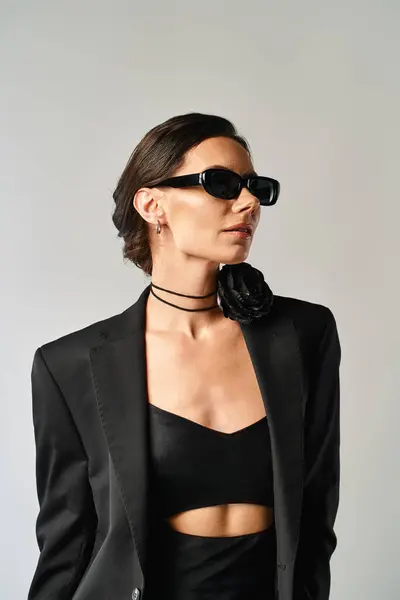Una donna alla moda trasuda fiducia in un abito nero e occhiali da sole, colpendo una posa in uno studio su uno sfondo grigio. — Foto stock