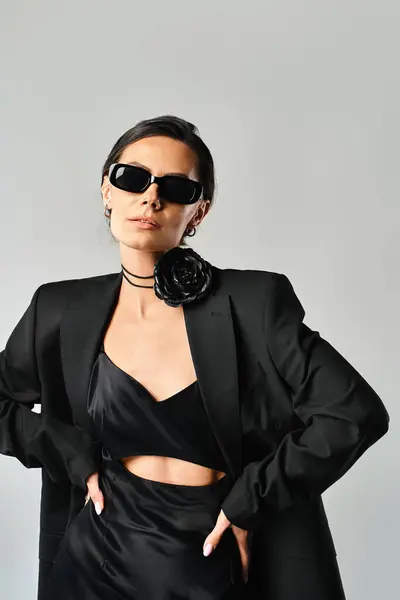 A fashionable woman exuding confidence in a black suit and chic sunglasses against a grey studio background. — Stock Photo