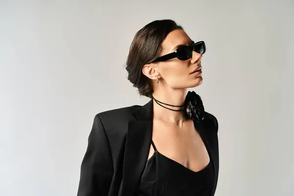 Eine stilvolle und geheimnisvolle Frau trägt in einem Studio vor neutralem grauen Hintergrund einen eleganten schwarzen Anzug und eine trendige Sonnenbrille. — Stockfoto