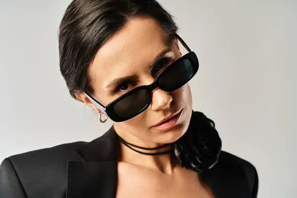 Una mujer con estilo emana confianza en un traje negro y gafas de sol de moda contra un fondo gris del estudio. - foto de stock