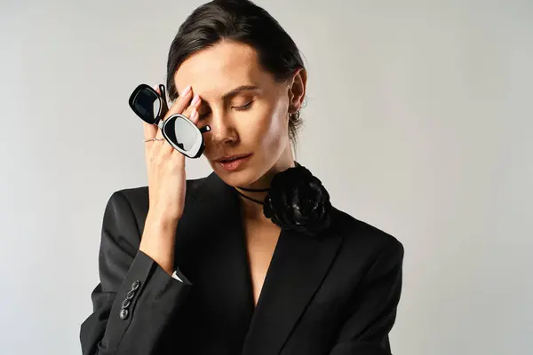 Una mujer de moda con un traje negro sostiene con confianza un par de gafas contra un fondo gris. - foto de stock