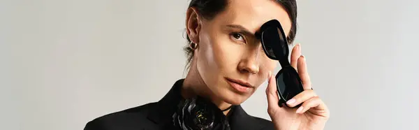 Une femme élégante en costume tient en toute confiance une paire de lunettes de soleil noires dans un cadre studio sur gris — Photo de stock