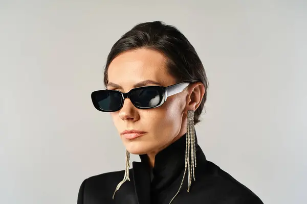 Una mujer de moda con gafas de sol posa en una chaqueta negra sobre un fondo gris del estudio. - foto de stock