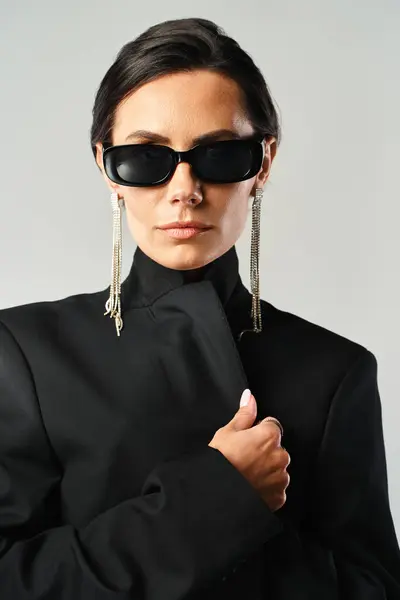 Une femme élégante avec des lunettes de soleil et une veste noire pose en toute confiance dans un studio sur un fond gris. — Photo de stock