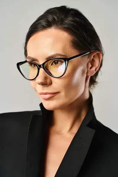Une femme élégante dans un blazer noir et des lunettes respire l'élégance dans un cadre studio sur un fond gris. — Photo de stock