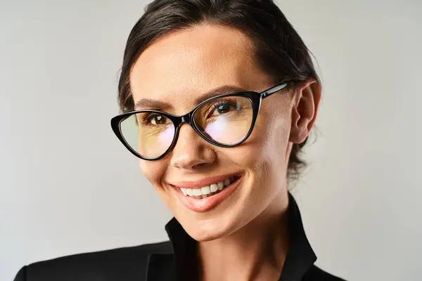 Una mujer elegante con gafas elegantes sonriendo brillantemente en un ambiente de estudio sobre un fondo gris. - foto de stock
