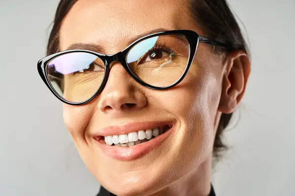 Una mujer de moda, con gafas, sonríe brillantemente a la cámara en un estudio sobre un fondo gris. - foto de stock