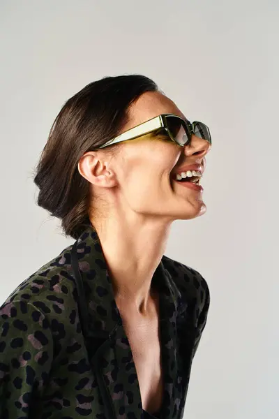 Una mujer elegante muestra con confianza una camisa estampada de leopardo y gafas de sol de moda en un entorno de estudio sobre un fondo gris. - foto de stock