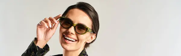 Uma mulher elegante exala confiança, balançando óculos de sol chiques e piscando um sorriso radiante em um estúdio contra um pano de fundo cinza. — Fotografia de Stock