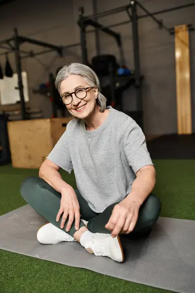 Joyful mature woman with glasses sitting with crossed legs on floor in gym and smiling at camera — Stock Photo
