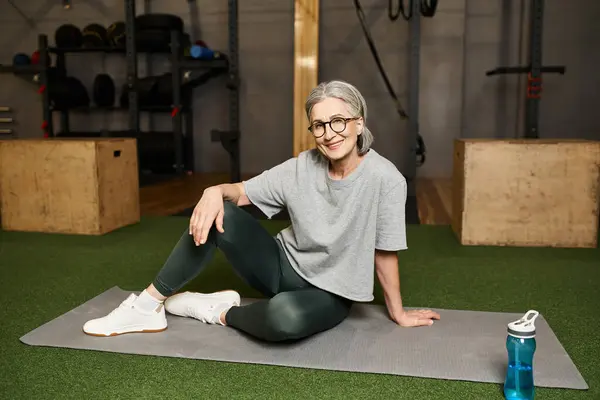 Positive beautiful mature woman in sportswear with glasses sitting on floor and smiling at camera — Stock Photo