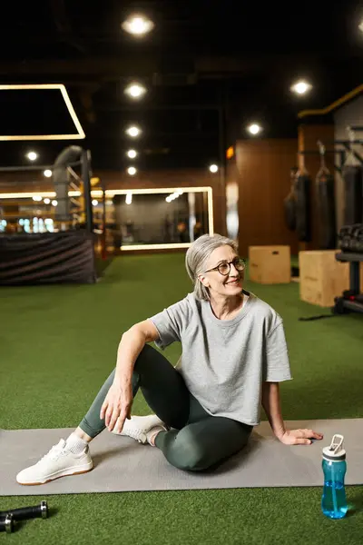 Séduisante femme mûre heureuse avec des cheveux gris et des lunettes assis sur le sol dans la salle de gym et regardant loin — Photo de stock
