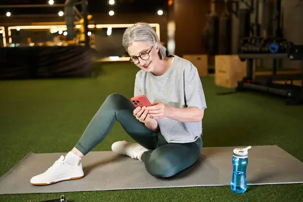 Good looking positive woman in cozy sportswear sitting in gym and looking at her smartphone — Stock Photo