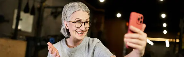 Positive senior sportswoman in comfy attire with glasses having video call while in gym, banner — Stock Photo