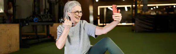 Jolly mature sportswoman in gray t shirt with glasses taking selfies while holding dumbbells, banner — Stock Photo