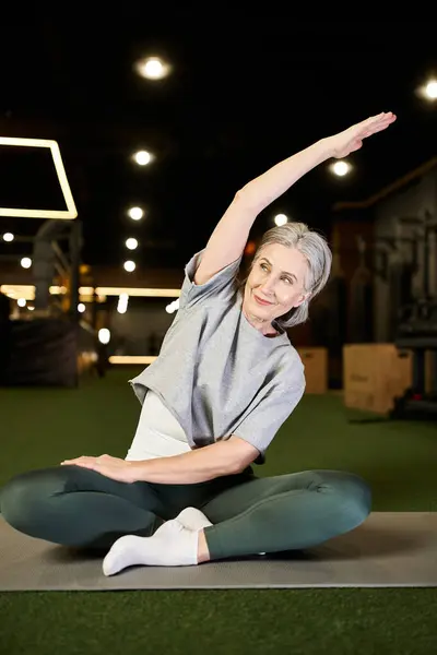 Positive beautiful mature sportswoman in comfy attire sitting and stretching her muscles in gym — Stock Photo