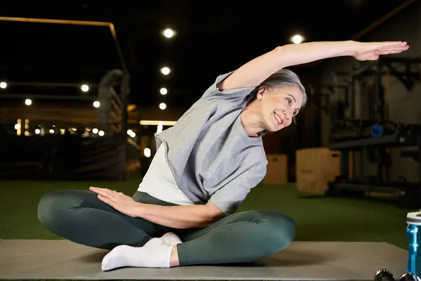 Cheerful appealing mature sportswoman in cozy attire sitting and stretching her muscles in gym — Stock Photo