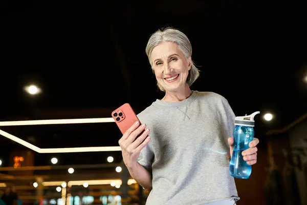 Contented mature sportswoman with gray hair holding water bottle and phone and smiling at camera — Stock Photo
