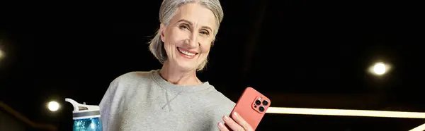 Mujer madura alegre con pelo gris sosteniendo botella de agua y teléfono y sonriendo a la cámara, pancarta - foto de stock