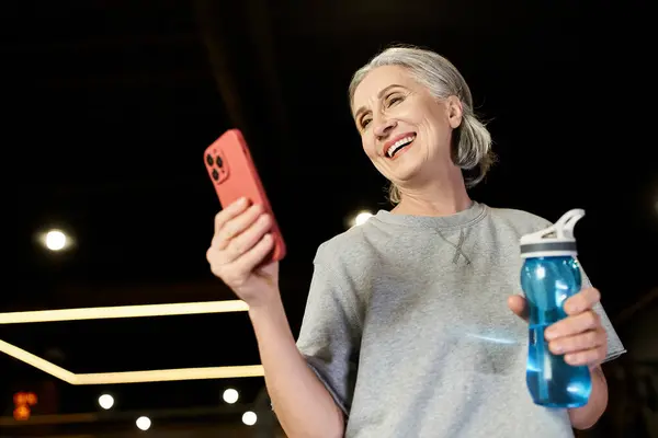 Positive beautiful senior woman in sportswear looking at her smartphone and holding water bottle — Stock Photo