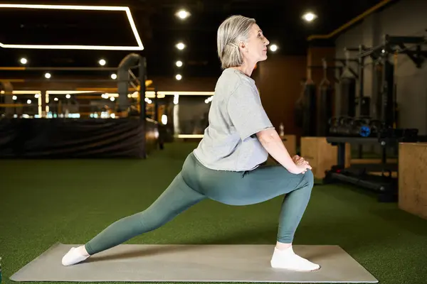 Séduisante sportive senior aux cheveux gris en tenue confortable étirant ses muscles pendant qu'elle est en salle de gym — Photo de stock