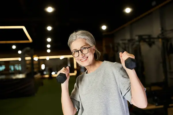 Fröhliche reife Frau mit Brille und grauem Haar, die mit Hanteln trainiert und in die Kamera lächelt — Stockfoto