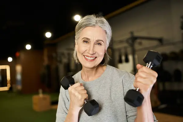 Bella donna anziana allegra in formazione abbigliamento sportivo attivamente con manubri e sorridente alla macchina fotografica — Foto stock