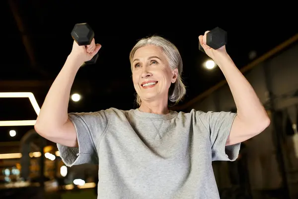 Attraente donna dai capelli grigi anziani in allenamento abbigliamento sportivo attivamente con manubri mentre in palestra — Foto stock