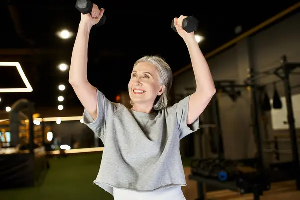 Beautiful mature gray haired woman in cozy sportswear training actively with dumbbells while in gym — Stock Photo