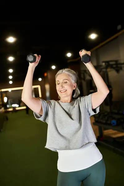 Belle femme mûre souriante dans des vêtements de sport confortables exerçant joyeusement avec des haltères dans la salle de gym — Photo de stock