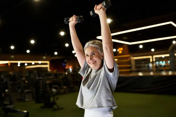Mature cheerful woman with gray hair in cozy attire training actively with dumbbells while in gym — Stock Photo