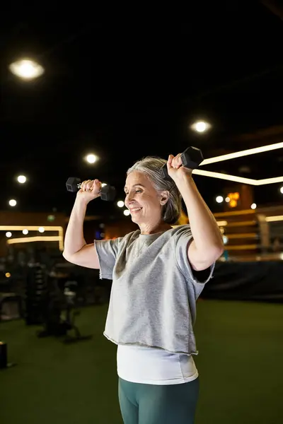Sporty senior woman in cozy attire training with dumbbells and smiling happily while in gym — Stock Photo