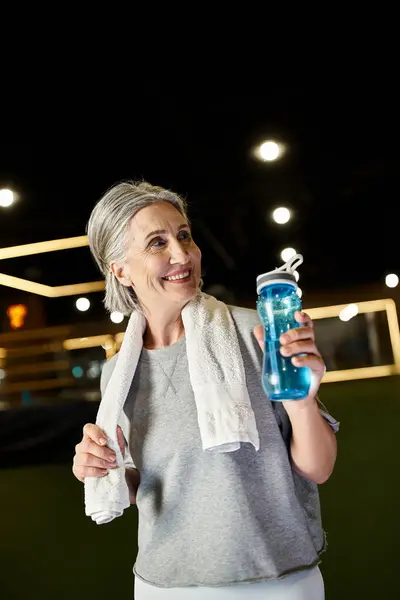 Good looking senior sporty woman with towel on shoulders holding water bottle and looking away — Stock Photo