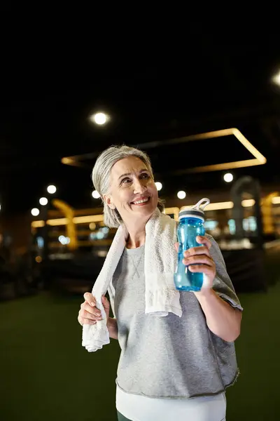 Belle femme sportive mature avec serviette sur les épaules tenant la bouteille d'eau et regardant loin — Photo de stock