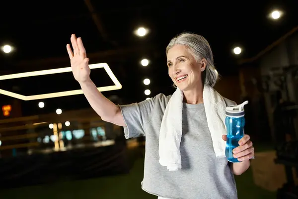 Pretty jolly mature sportswoman with towel on shoulders and water bottle in hands waving actively — Stock Photo