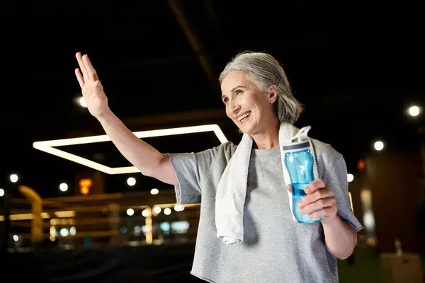 Good looking jolly senior sportswoman with towel on shoulders and water bottle waving actively — Stock Photo