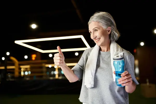 Merry athletic mature woman in sportswear with towel on shoulders with water bottle showing thumb up — Stock Photo